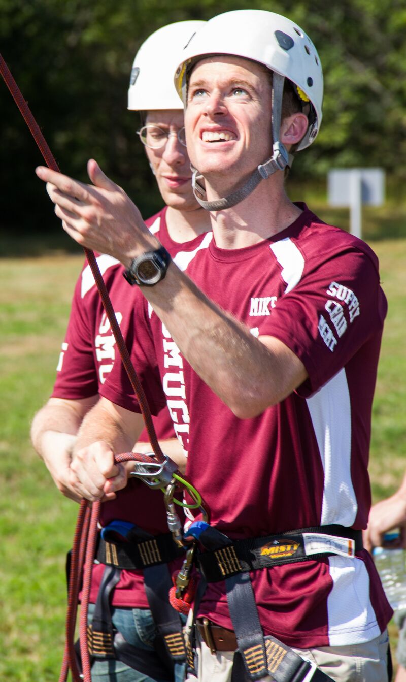 The image shows two men wearing helmets and maroon shirts, likely engaged in an outdoor activity like rock climbing or rappelling. They are holding ropes and appear focused, with one looking upwards. The setting seems to be a sunny day, with green grass visible in the background.